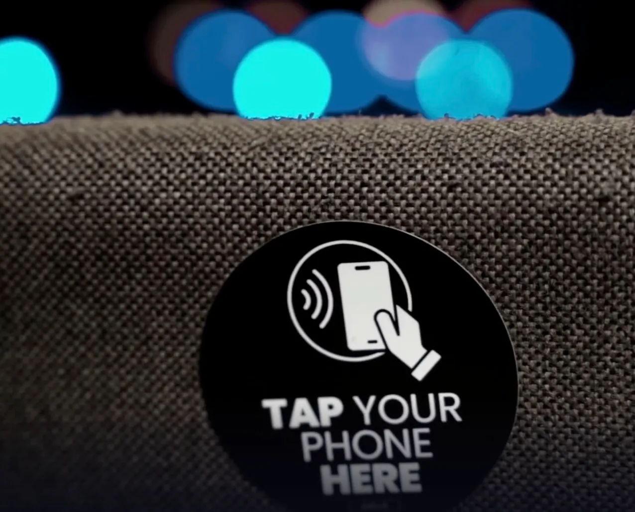 A round Tap Your Phone Here NFC tag attached to an upholstered chair